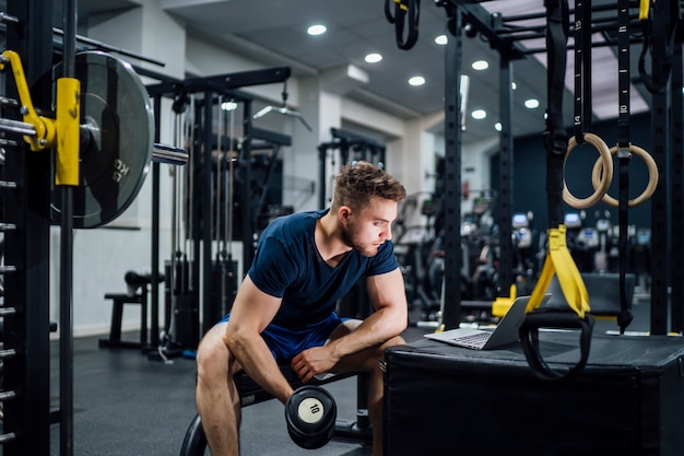 Przystojny mężczyzna ćwiczy bodybuilding obciąża w gym i ogląda środki na laptopie