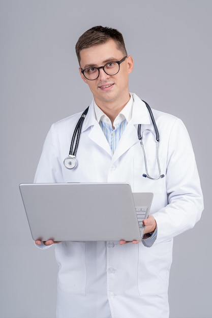 Przystojny mężczyzna medyk stoi z laptopem w rękach. Uśmiechnięty lekarz czyta dobre wieści na ekranie.