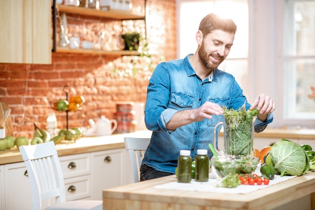 Przystojny mężczyzna napełnia mikser zielonym surowym jedzeniem, przygotowując wegetariański koktajl w kuchni w domu