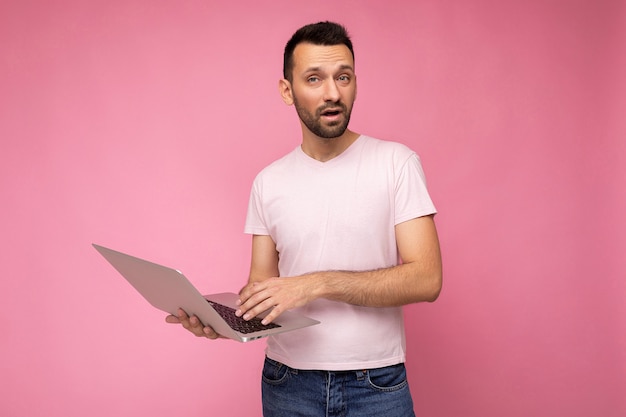 Przystojny mężczyzna pyta bruneta, trzymając laptopa i pokazując rękę patrząc na kamery w t-shirt na na białym tle różowym.