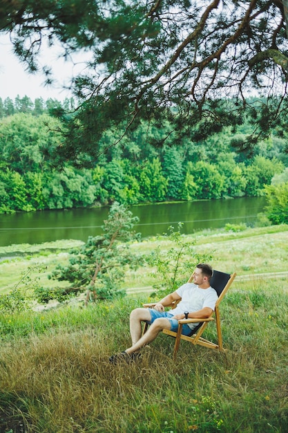 Przystojny mężczyzna siedzi na drewnianym krześle w naturze Młody przystojny facet siedzi na ławce w cieniu drzew i cieszy się otaczającą przyrodą w słoneczny dzień