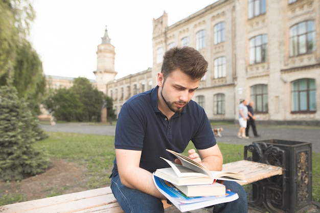 Przystojny mężczyzna siedzi na ławce w kampusie uniwersyteckim i czyta książkę.