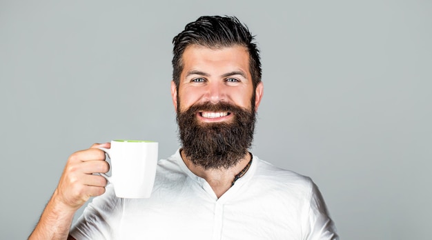 Przystojny mężczyzna trzyma filiżankę kawy, herbaty. Brodaty mężczyzna uśmiechający się pokazując znak ok. Dzień dobry, człowieku herbatka, ok. Uśmiechnięty mężczyzna hipster z filiżanką świeżej kawy, szczęśliwy człowiek pokazując znak ok. Koncepcja poranna