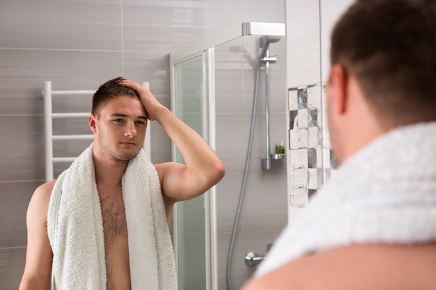 Przystojny Mężczyzna Trzymający Ręcznik Na Ramionach Po Zabiegach Mycia, Patrząc W Lustro I Stojąc W Nowoczesnej, Wyłożonej Kafelkami łazience