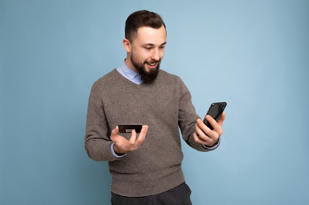 Przystojny mężczyzna ubrany w codzienne ubrania na białym tle na tle ściany trzymając i za pomocą telefonu i karty kredytowej dokonywanie płatności patrząc na ekran smartfona.