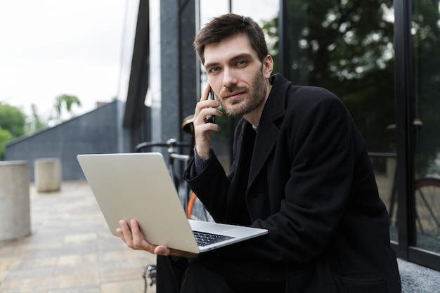 Przystojny mężczyzna ubrany w płaszcz siedzi na ulicy, używając laptopa, rozmawiając przez telefon komórkowy