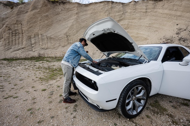Przystojny mężczyzna w dżinsowej kurtce i czapce stoi w pobliżu swojego białego samochodu typu muscle car z otwieraną maską, sprawdzając poziom oleju w silniku
