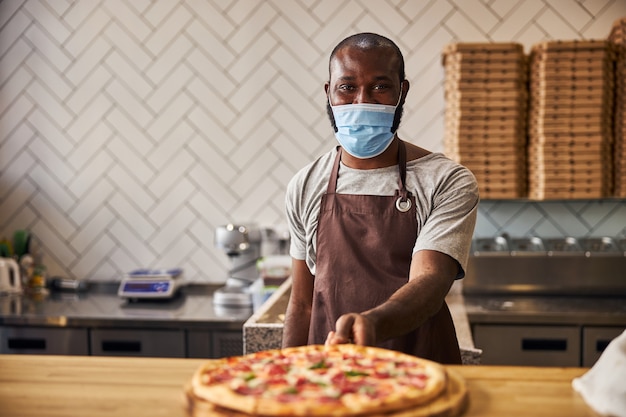 Przystojny mężczyzna w masce ochronnej stojący przy ladzie ze świeżo upieczoną pizzą w pizzerii
