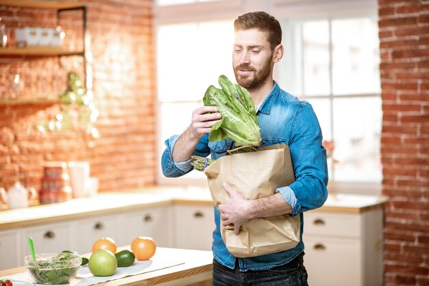 Przystojny mężczyzna w niebieskiej koszuli rozpakowujący zdrową żywność z torby na zakupy w kuchni