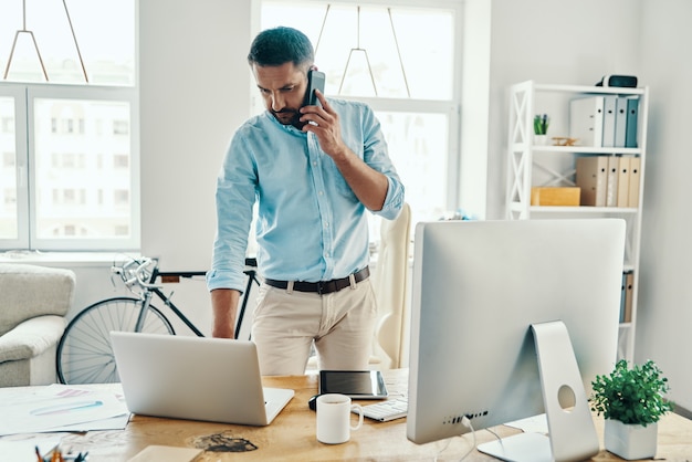 Przystojny mężczyzna w średnim wieku w eleganckim stroju casual rozmawia przez telefon stojąc w biurze