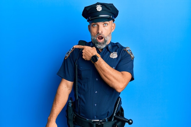 Przystojny mężczyzna w średnim wieku w mundurze policyjnym zaskoczony, wskazując palcem na bok, z otwartymi ustami zdumiony wyraz.