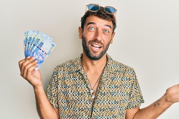 Przystojny mężczyzna z brodą trzymający banknoty 100 randów południowoafrykańskich świętujący osiągnięcie z radosnym uśmiechem i wyrazem twarzy zwycięzcy z podniesioną ręką