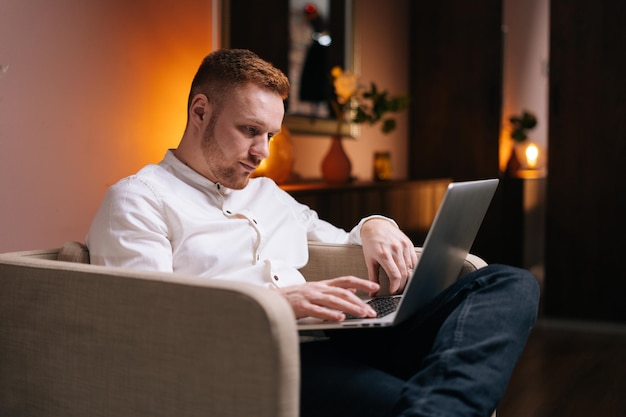 Przystojny mężczyzna za pomocą laptopa siedząc w miękkim fotelu w domu w przytulnym salonie. Koncepcja pracy zdalnej w domu podczas pandemii koronawirusa COVID-19.