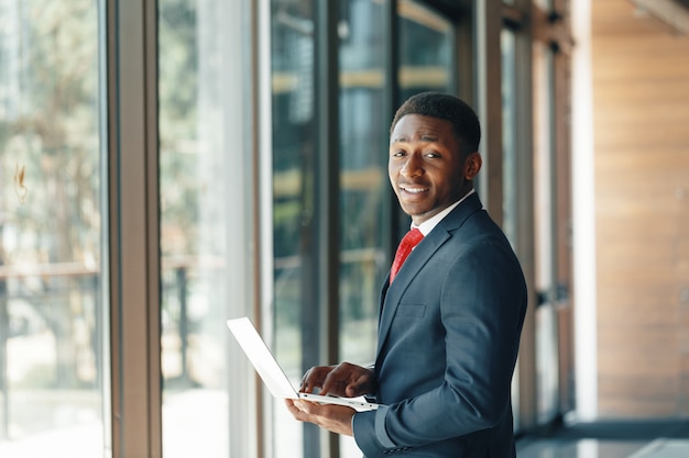 Przystojny młody Afro amerykański biznesmen w klasycznym garniturze trzyma laptopa i uśmiecha się