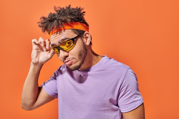 przystojny młody Afroamerykanin dostosowuje okulary przeciwsłoneczne i patrzy na kamerę na pomarańczowym tle