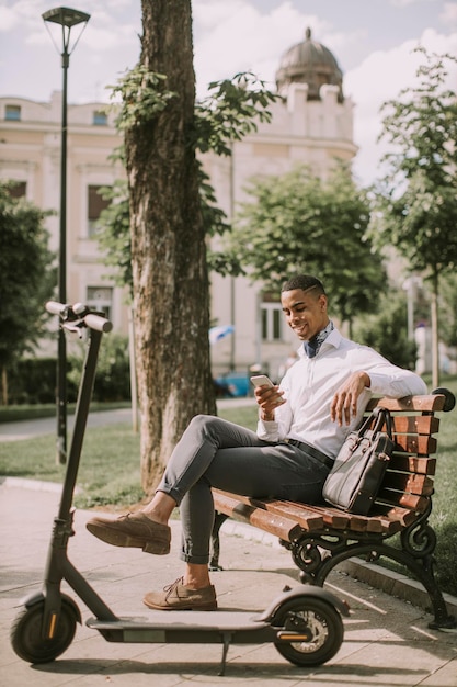 Przystojny Młody Afroamerykanin Korzystający Z Telefonu Komórkowego Siedząc Na ławce Przy Skuterze Elektrycznym