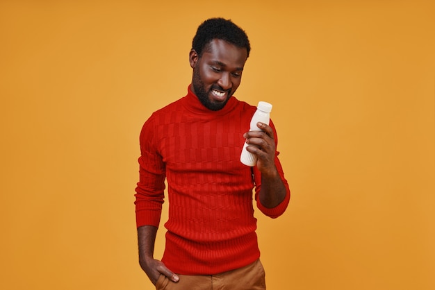 Przystojny młody afrykański mężczyzna patrzący na butelkę i uśmiechający się stojąc na żółtym tle