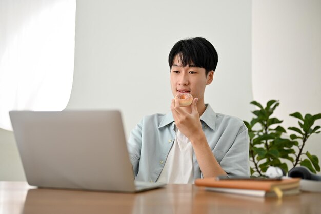 Przystojny młody azjatycki student jedzący pączki podczas pracy nad swoim projektem na laptopie