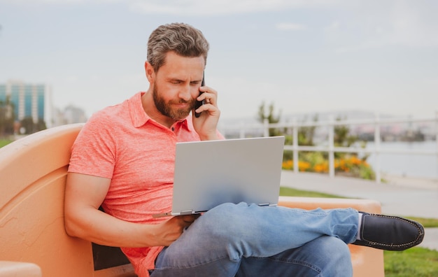 Przystojny młody biznesmen pracujący z laptopem na zewnątrz rozmawia przez telefon komórkowy