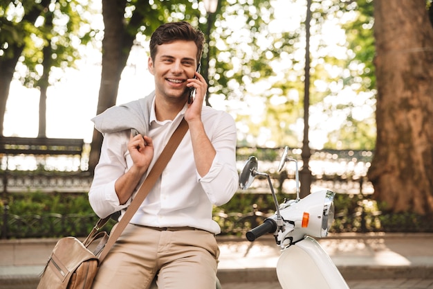 Przystojny młody biznesmen siedzi na motocyklu na zewnątrz, rozmawia przez telefon komórkowy