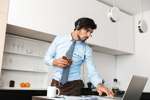 Przystojny młody biznesmen w kuchni zjeść kanapkę przy użyciu komputera przenośnego na sobie słuchawki.