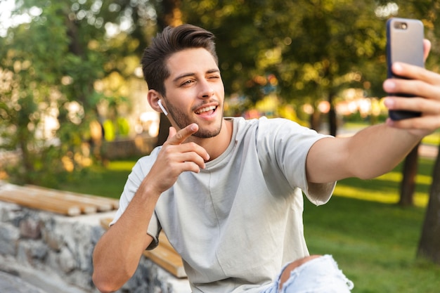 Przystojny, młody chłopak siedzi w parku na świeżym powietrzu, biorąc selfie przez telefon komórkowy i słuchając muzyki przez słuchawki