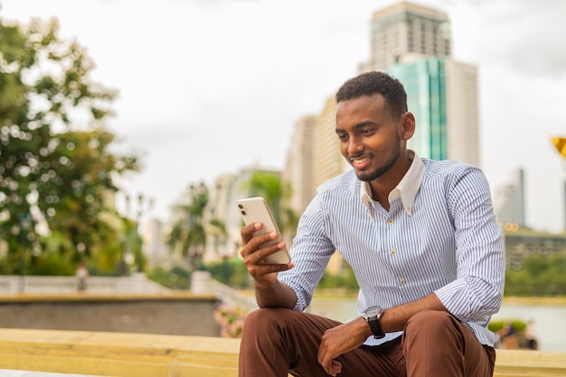 Przystojny młody czarny biznesmen w parku przy użyciu telefonu komórkowego