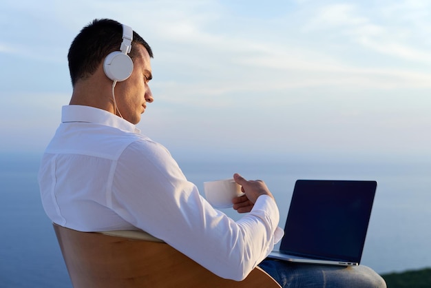 przystojny młody człowiek relaksujący się i pracujący na laptopie na balkonie w domu, patrząc na zachód słońca