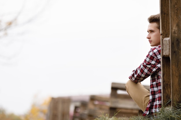 Przystojny Młody Facet W Koszuli W Kratę, Siedząc Na Trawie W Pobliżu Drewnianego Ogrodzenia W Jesienny Dzień