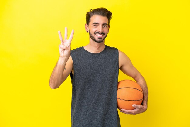 Przystojny młody koszykarz na białym tle na żółtym tle szczęśliwy i liczący trzy palcami