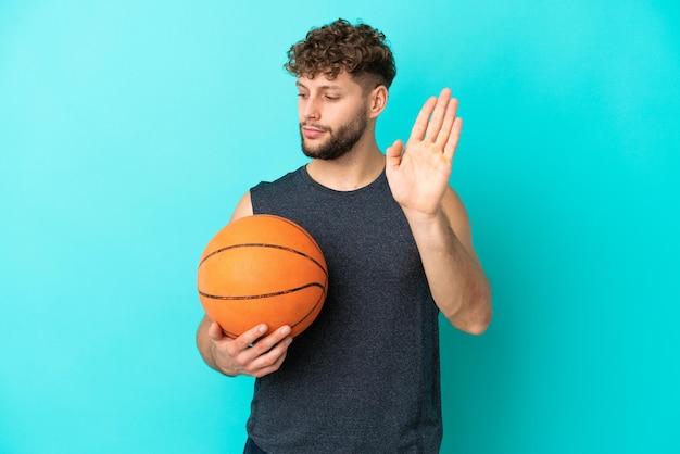Przystojny młody mężczyzna grający w koszykówkę na białym tle na niebieskim tle, wykonując gest zatrzymania i rozczarowany