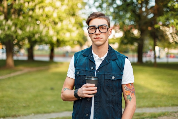 Zdjęcie przystojny młody mężczyzna hipster z okularami i kawą spacerując w słoneczny dzień w parku