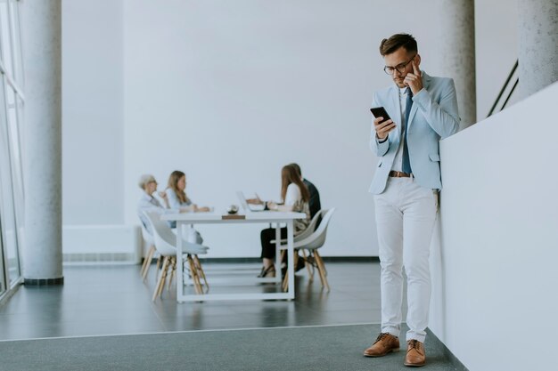 Przystojny młody mężczyzna korzystający z telefonu komórkowego w biurze, podczas gdy jego koledzy pracują w tle