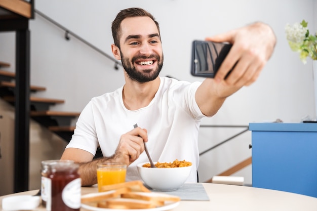 Przystojny Młody Mężczyzna O śniadanie Siedząc W Kuchni, Biorąc Selfie