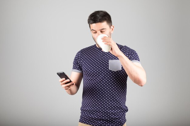 Przystojny młody mężczyzna pije kawę i używa swojego smartfona odizolowanego na szaro