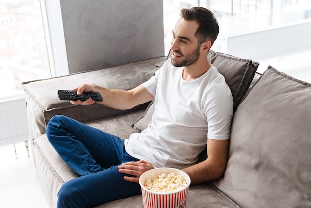 Przystojny Młody Mężczyzna Siedzi Na Kanapie W Domu, Je Popcorn, Ogląda Telewizję