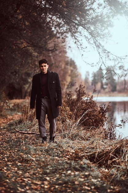Przystojny Młody Mężczyzna Ubrany W Czarny Płaszcz, Spacerujący W Pobliżu Jeziora W Jesiennym Lesie.