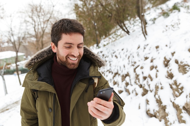 Przystojny Młody Mężczyzna Ubrany W Kurtkę Zimową Stojący Na Zewnątrz, Przy Użyciu Telefonu Komórkowego