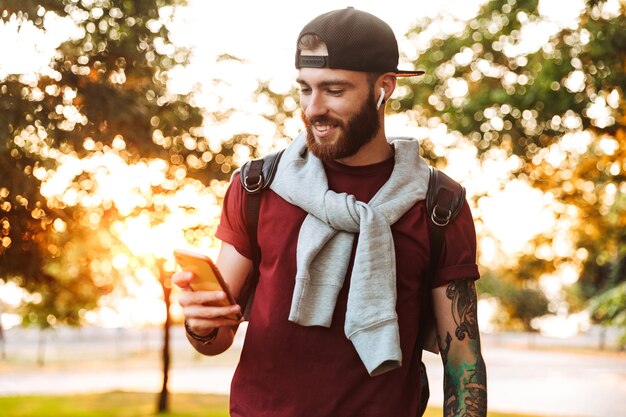 Przystojny młody mężczyzna ubrany w zwykłe ubranie spacerujący po parku, słuchający muzyki przez bezprzewodowe słuchawki, używający telefonu komórkowego
