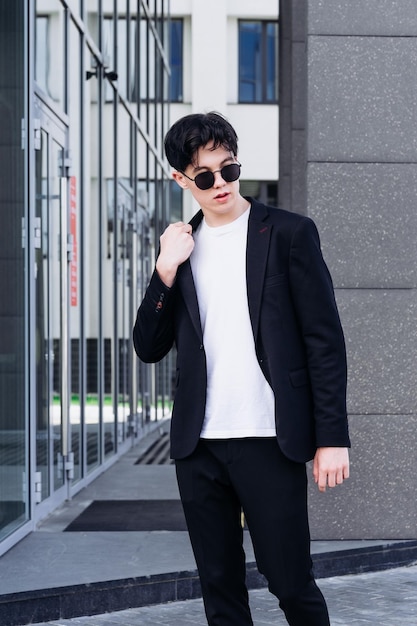 Przystojny młody mężczyzna w czarnej koszuli stojący na tle miejskich budynków i odwracający wzrok przez czarne okulary przeciwsłoneczne