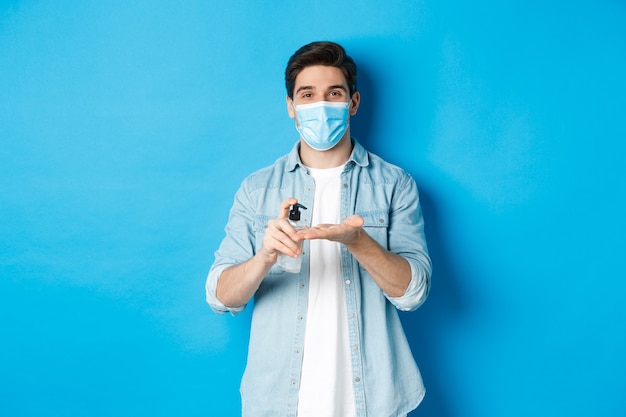 Przystojny młody mężczyzna w masce medycznej dezynfekuje ręce środkiem odkażającym, stosując środek antyseptyczny, stojąc przed niebieską ścianą