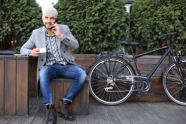 Przystojny Młody Mężczyzna W Szarym Płaszczu I Kapeluszu Rozmawia Przez Telefon Komórkowy I Uśmiecha Się Siedząc W Pobliżu Swojego Roweru Na Zewnątrz.