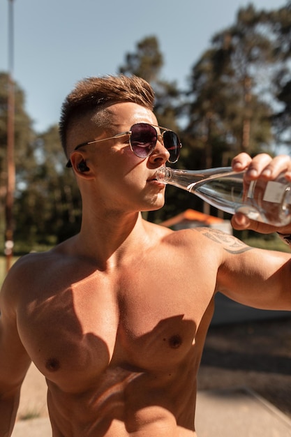 Przystojny młody mężczyzna z muskularnym ciałem ćwiczy i pije wodę na zewnątrz ze światłem słonecznym Zdrowy styl życia i koncepcja równowagi wodnej