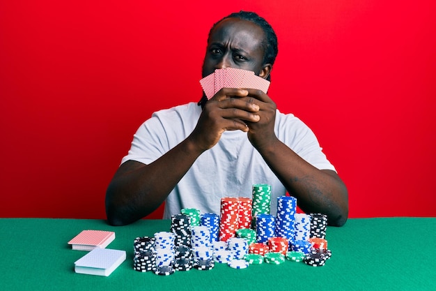 Przystojny młody murzyn grający w pokera hazardowego zakrywający twarz kartami w szoku, wyglądający sceptycznie i sarkastycznie zaskoczony otwartymi ustami