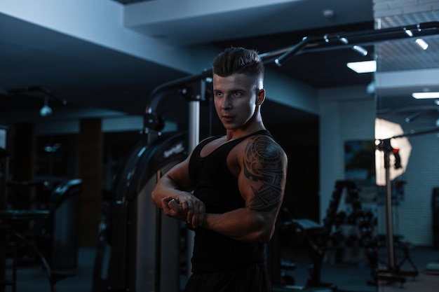 Przystojny młody sportowiec o muskularnym ciele z tatuażem w sportowej czarnej koszulce z szortami pokazuje mięśnie na siłowni