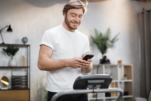 Przystojny młody sportowiec za pomocą telefonu patrząc na zajęcia fitness online podczas ćwiczeń