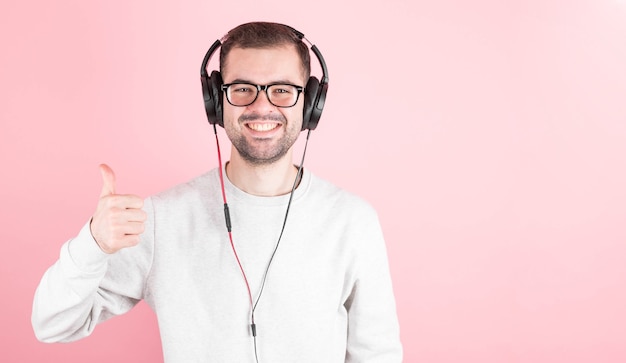 Przystojny młody stylowy mężczyzna w słuchawkach, słuchając dobrej muzyki, pokazując super znak, stojąc na różowej ścianie