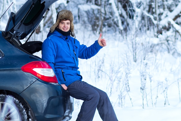 Zdjęcie przystojny młody szczęśliwy mężczyzna w ciepłych zimowych ubraniach siedzi na bagażniku samochodu w zimny śnieżny dzień