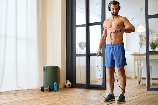 Przystojny Muskularny Mężczyzna Trzymający Skakankę I Sprawdzający Zużycie Kalorii Na Monitorze Fitness Podczas Słuchania Muzyki Przez Bezprzewodowe Słuchawki
