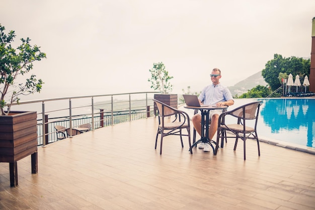 Przystojny, odnoszący sukcesy młody biznesmen siedzący przy stole przy basenie z laptopem z widokiem na Morze Śródziemne Praca zdalna na wakacjach Koncepcja wakacji
